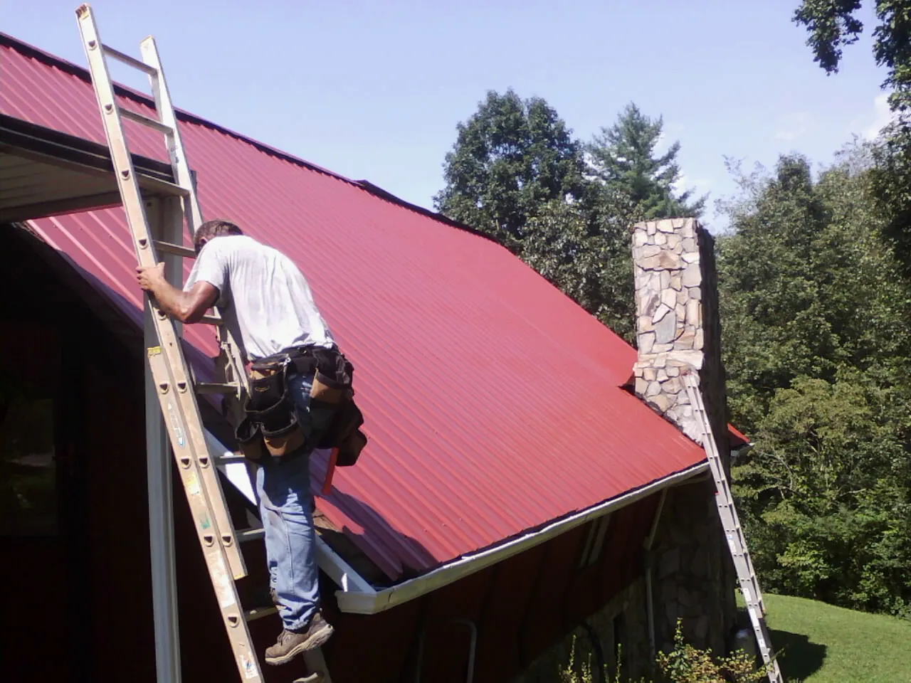 Roofer on a ladder beside a red metal roof and stone chimney — Soffit & Fascia Repair by Roofing Brothers in Cowpens
